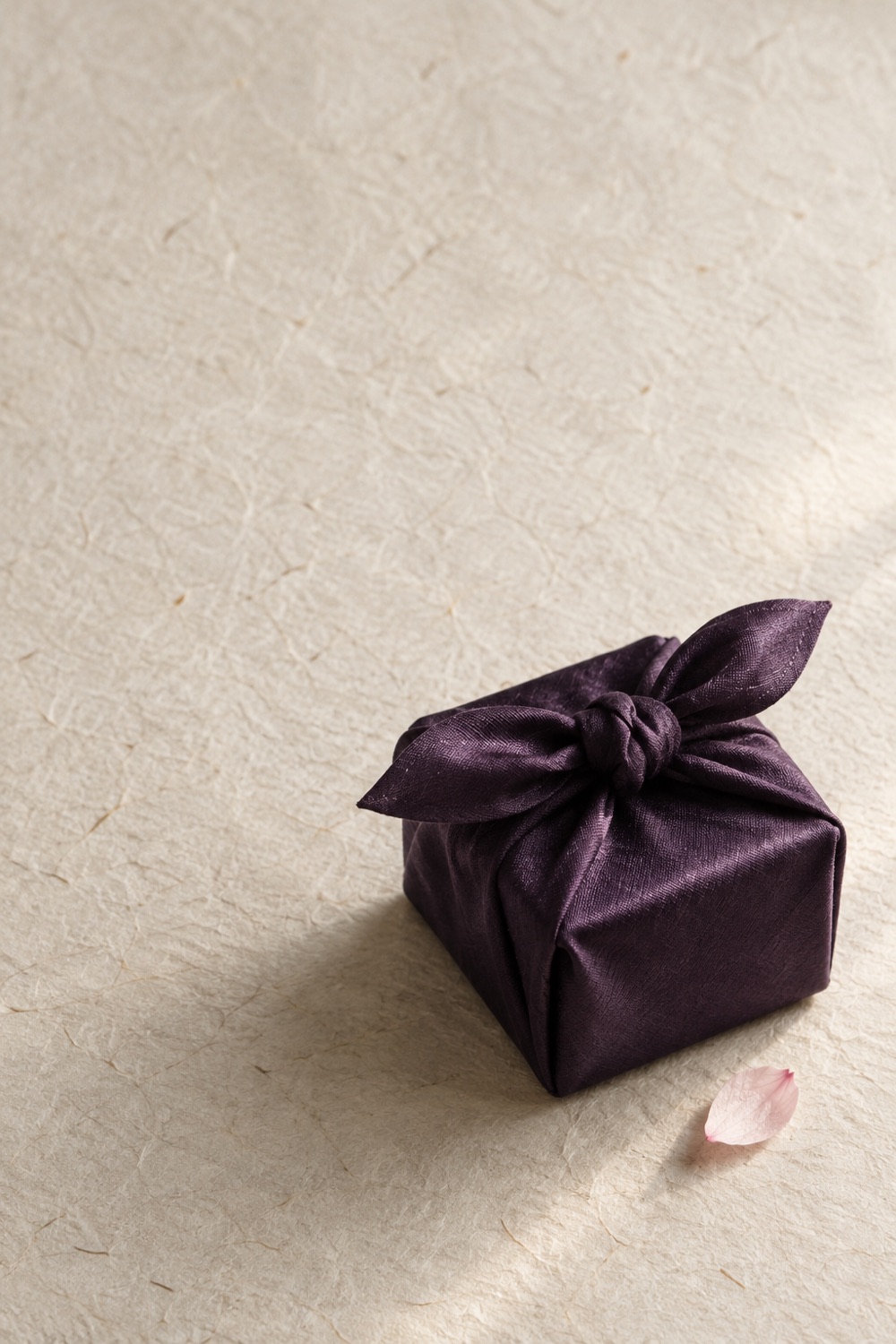 A small box wrapped in aubergine silk on washi paper, a cherry blossom petal beside it